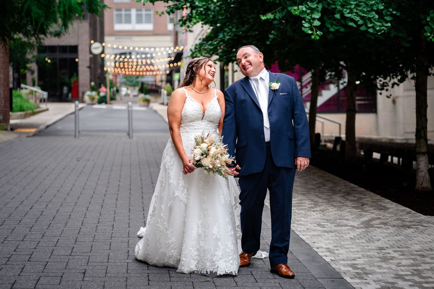 Wedding couple poses on brick pathway surrounded by lush greenery and white columns.