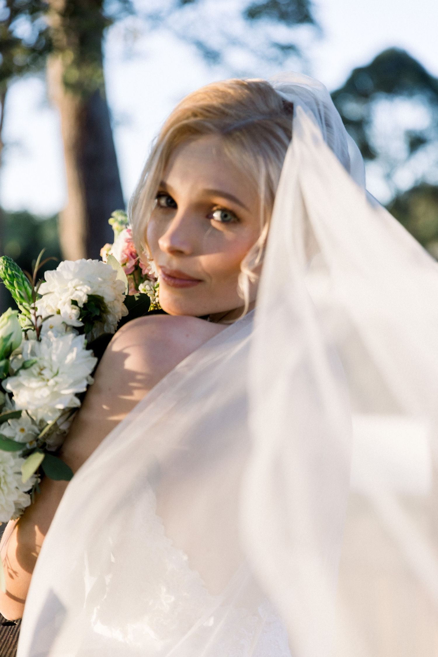 A bride in a flowing white veil holds a bouquet of white and pink flowers in an outdoor garden setting.