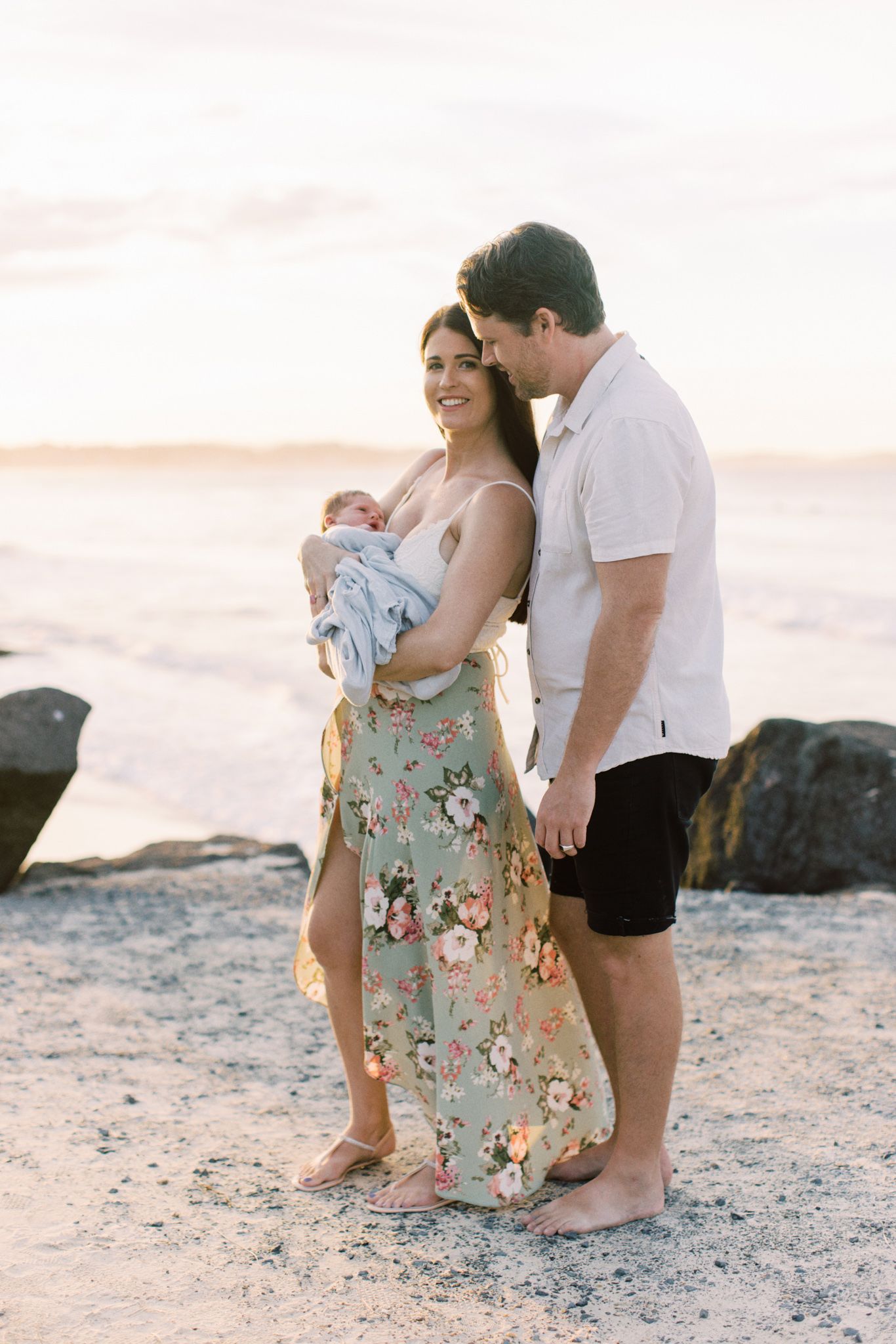 A couple stands on a beach at sunset with their newborn baby, the mother wearing a floral dress and father in white shirt.