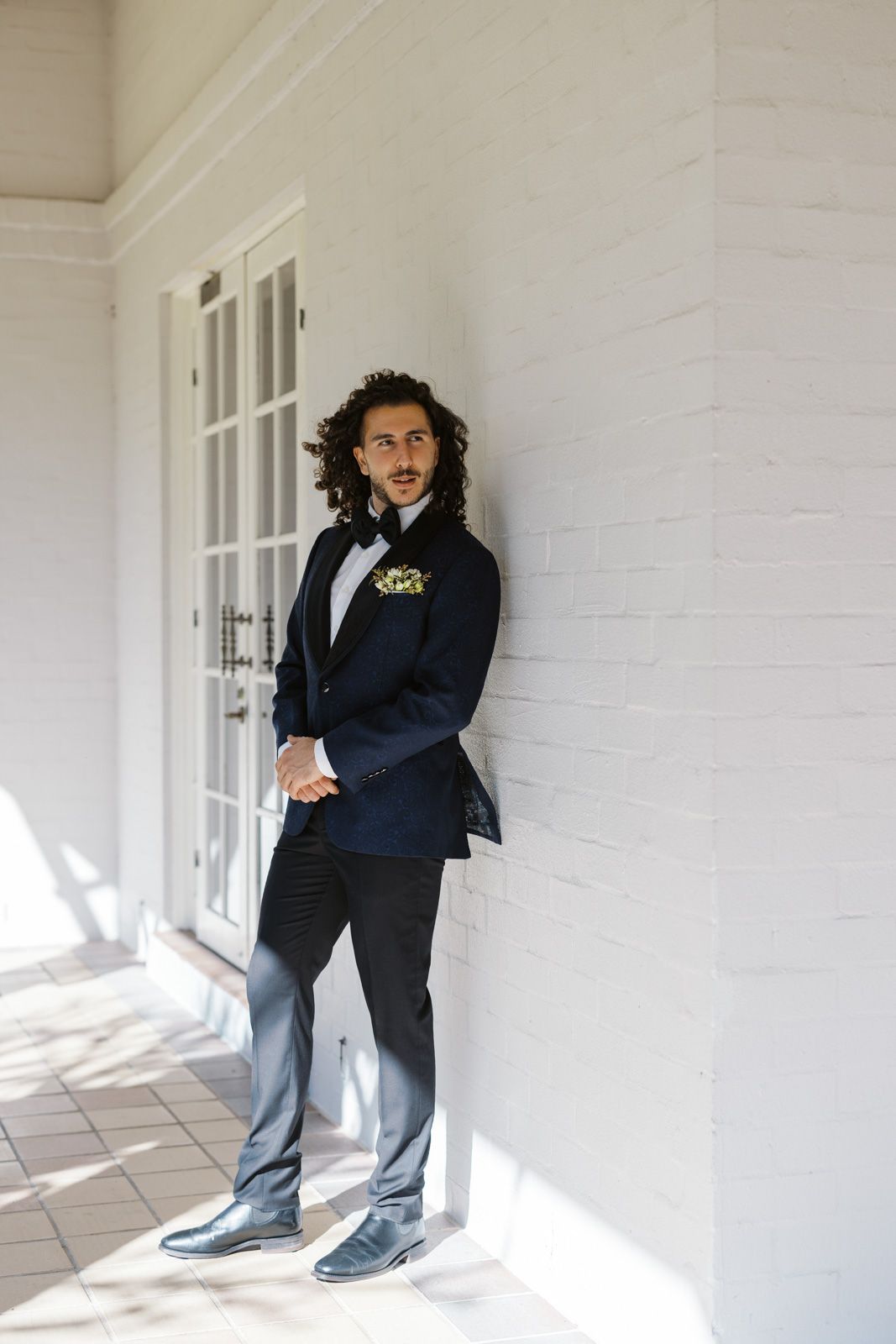 A person in a formal navy suit and bowtie stands confidently on a white porch in natural lighting.