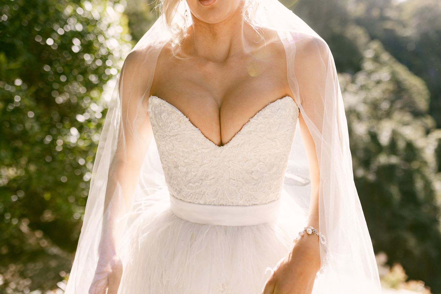 Bride in strapless lace wedding gown with flowing veil outdoors in natural sunlight.