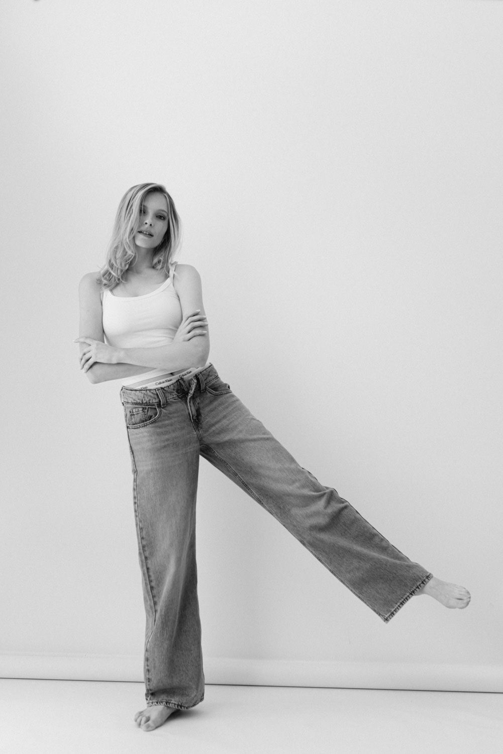 Black and white photo of a blonde woman in wide-leg jeans and white tank top posing with one leg raised.