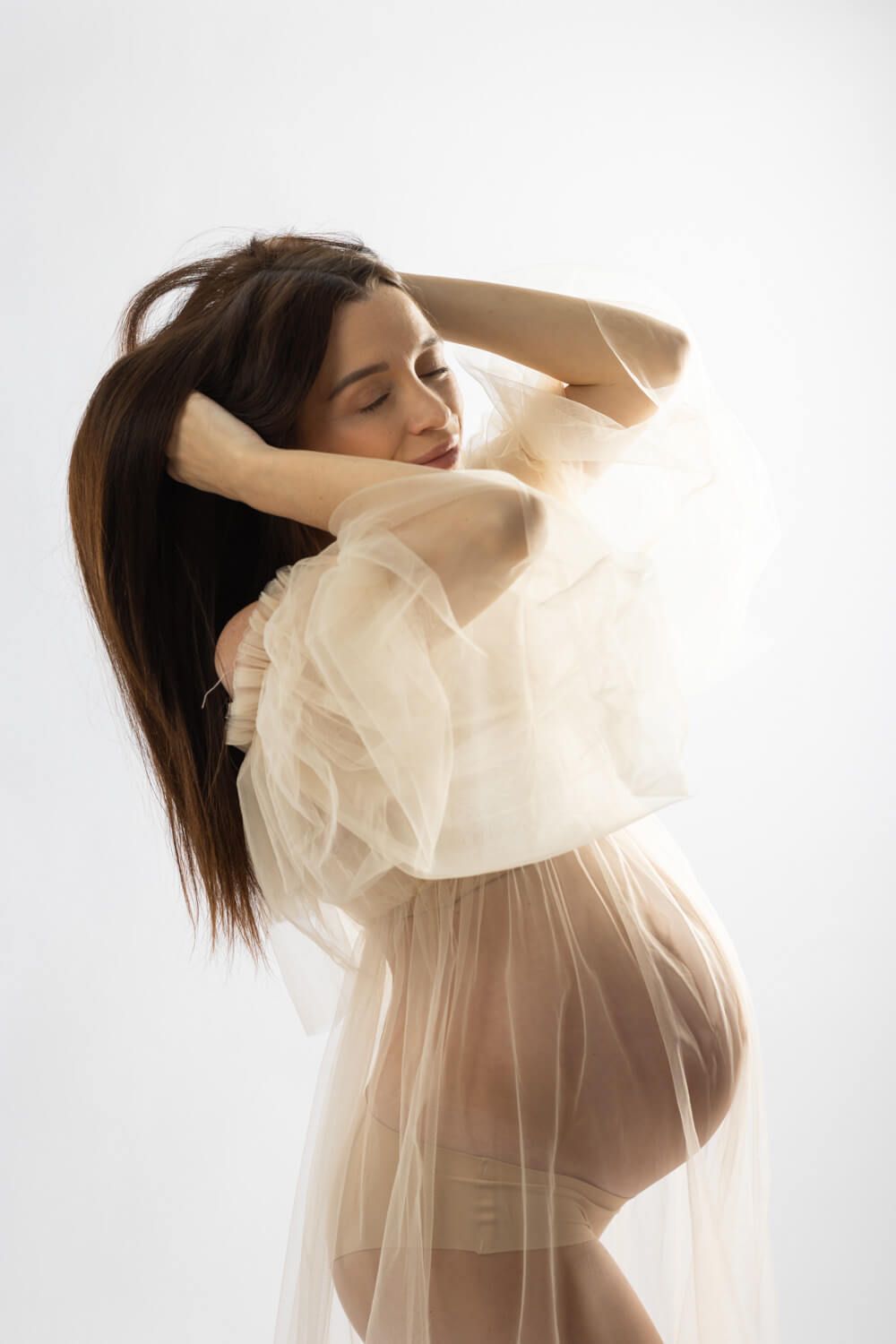 Pregnant woman in sheer white tulle robe posing gracefully with hands in hair against white background.