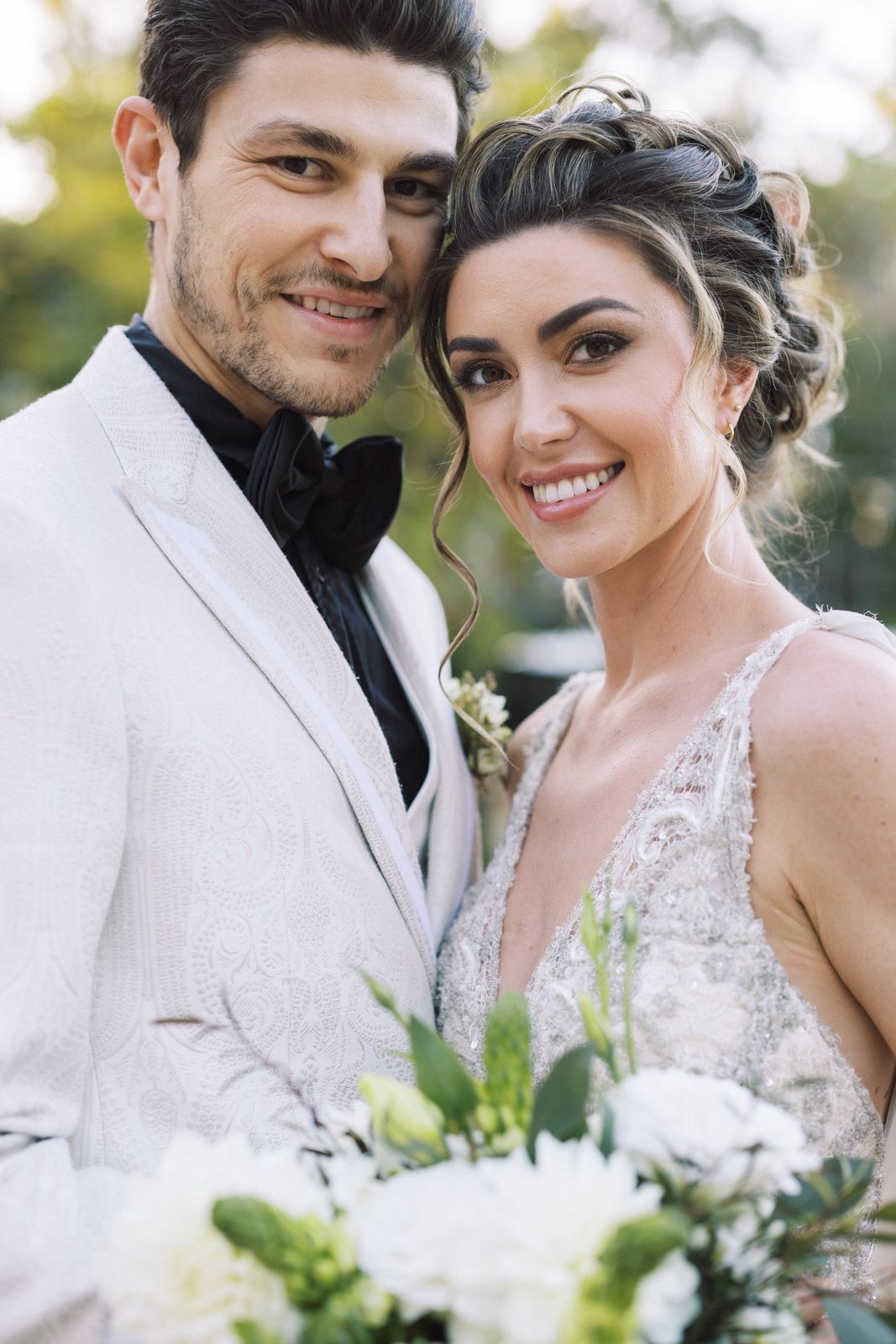 A happy couple poses together on their wedding day wearing white attire and holding a bouquet with greenery.