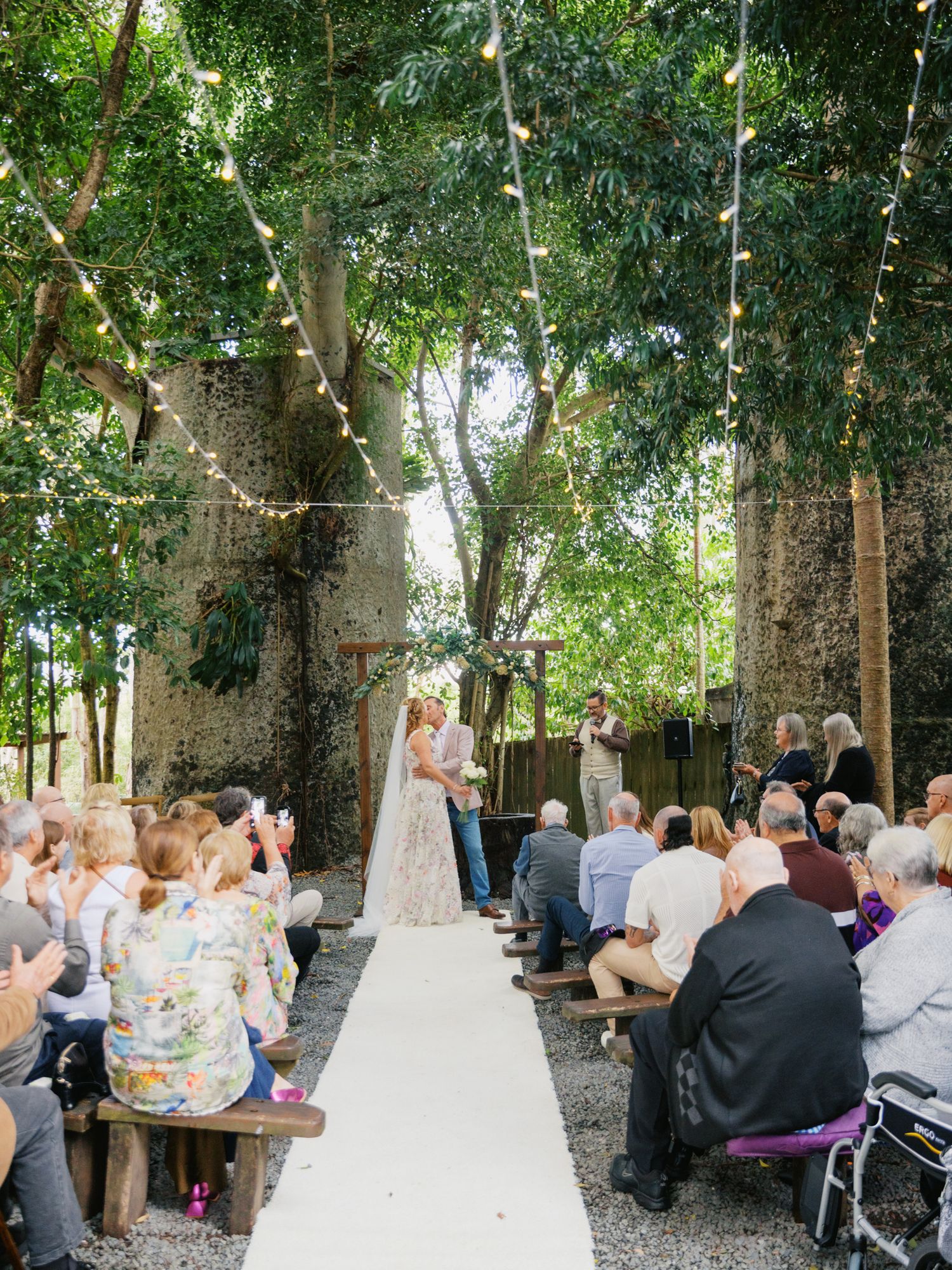 Outdoor wedding ceremony under string lights and lush trees, guests seated on wooden benches along white aisle at Fleay's Wedding Events.