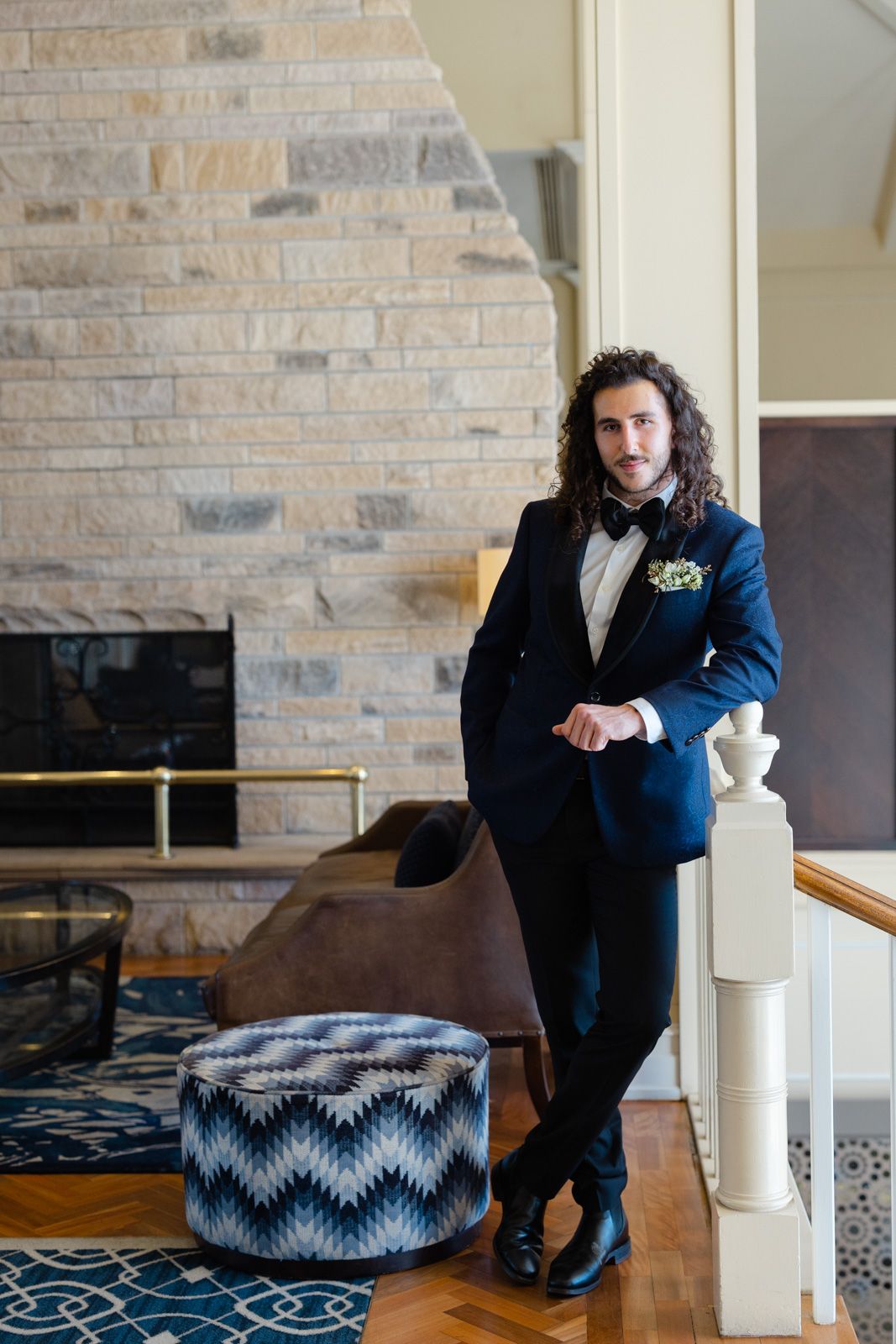 A person in a navy blue tuxedo and bow tie leans against a wall in an elegant room with patterned decor.