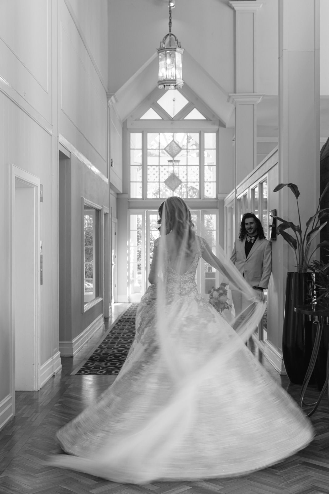 Black and white photo of a bride twirling in her wedding gown in an elegant hallway with a decorative window and lantern.