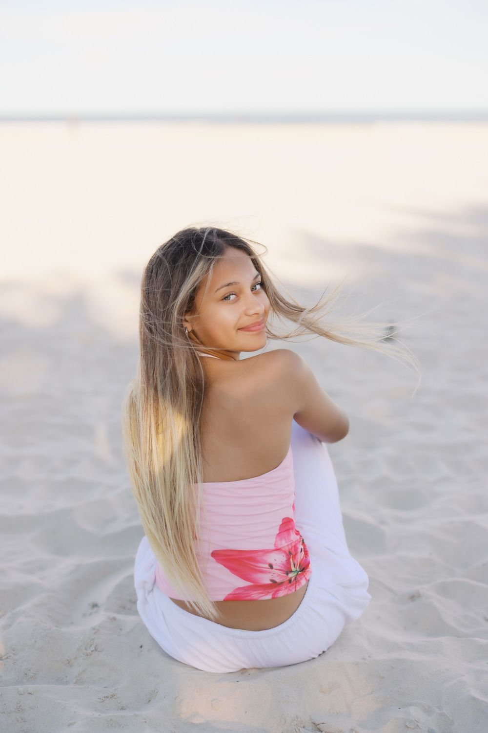 Young girl with long blonde ombre hair sits on sandy beach in pink floral top, smiling over her shoulder.