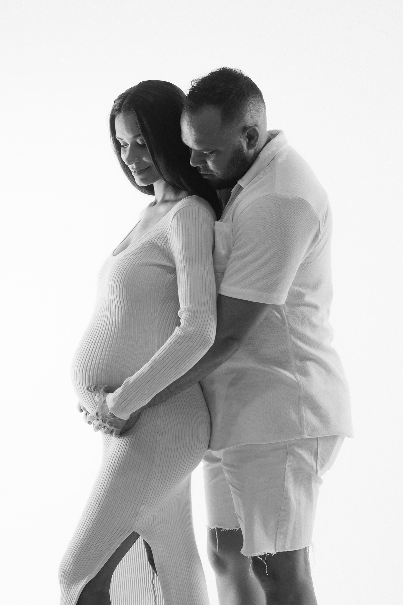 A tender maternity photoshoot with couple in white outfits embracing against light background.