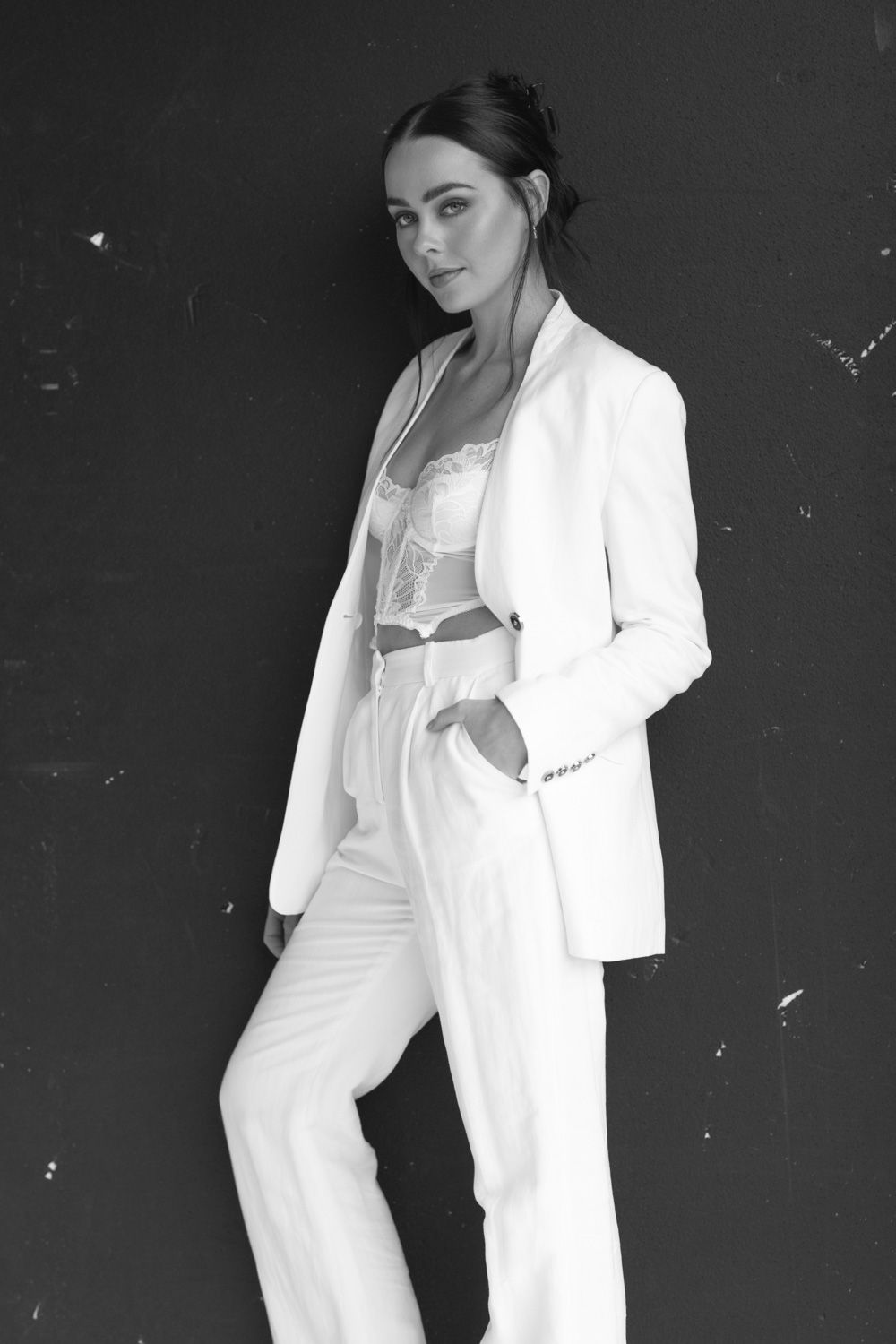 Fashion portrait in white pantsuit and lace top against dark background.