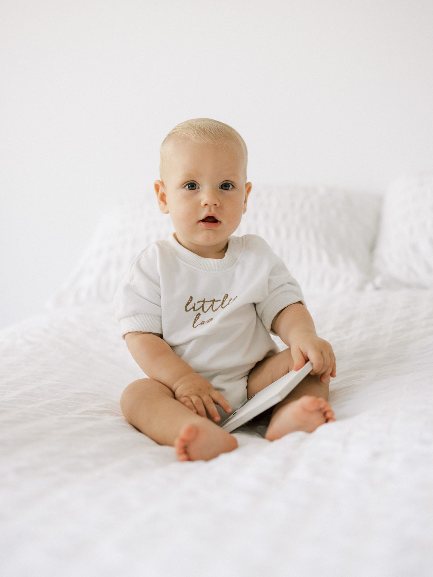 Baby in white onesie sitting on white bedding holding a device.