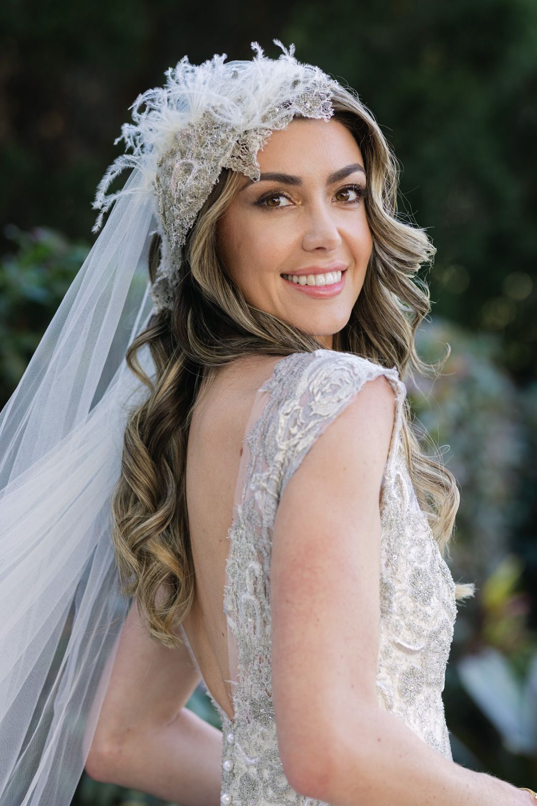 Beautiful bride wearing a lace wedding dress and delicate feathered headpiece with flowing veil smiles over her shoulder.