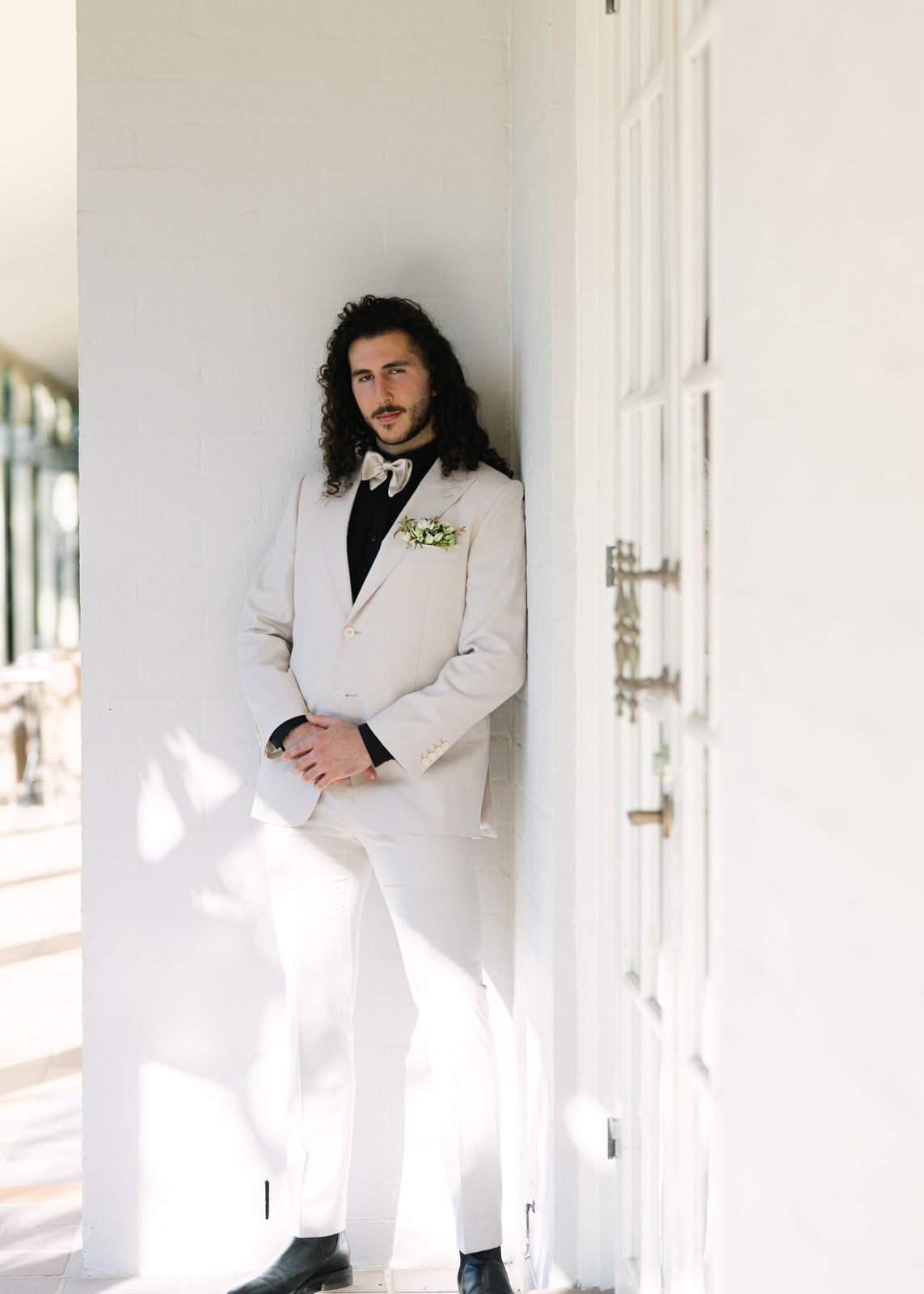 Person in a white suit leans against a bright white wall in an elegant interior hallway.