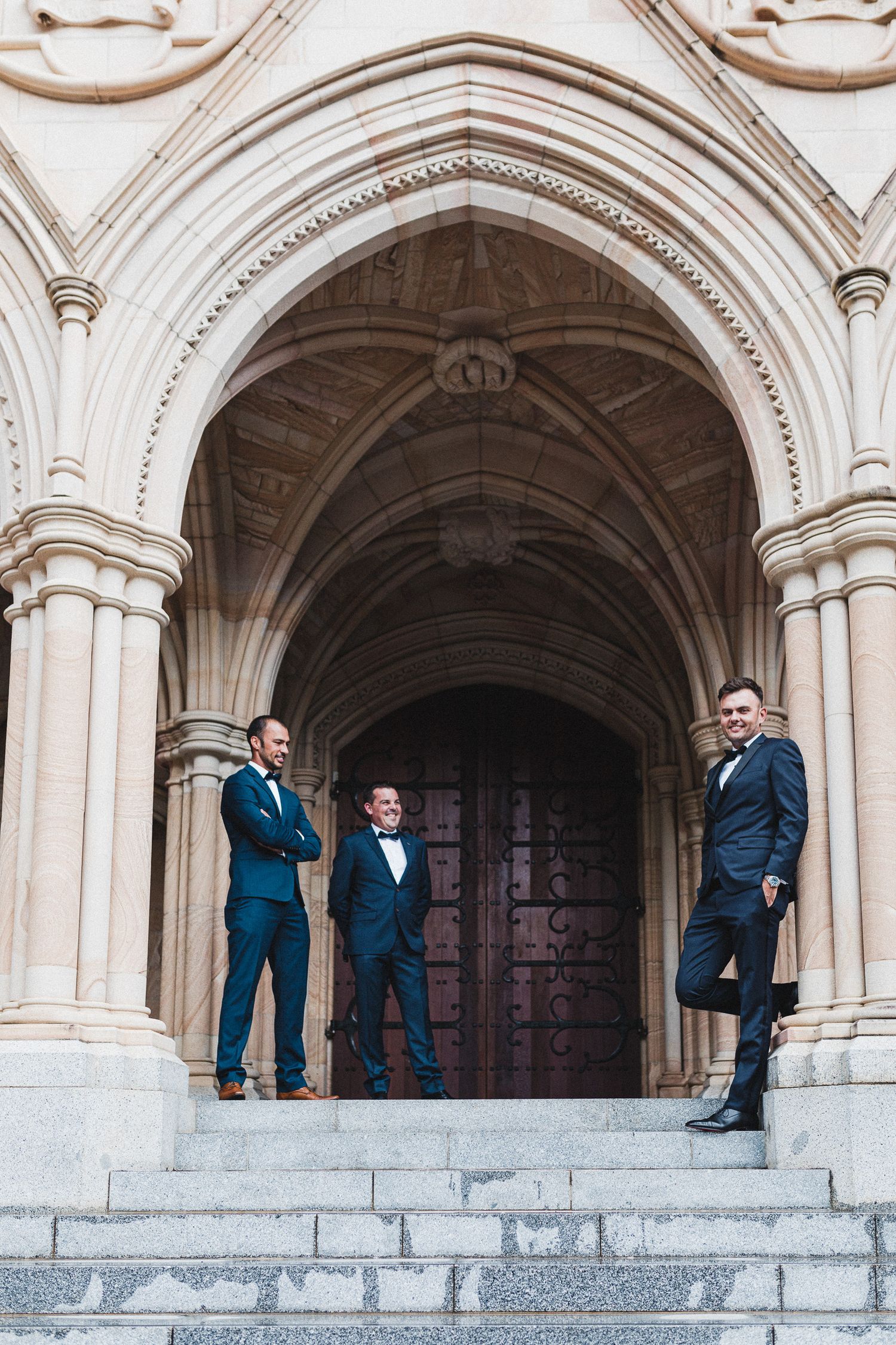 Groomsmen in navy suits posing on stone steps of historic Gothic architecture building.
