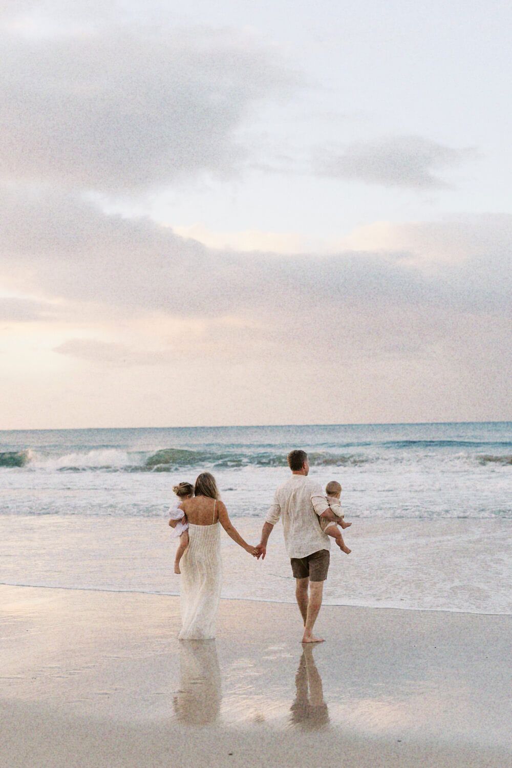 A family walks hand-in-hand along a serene beach at sunset, parents carrying young children toward gentle ocean waves.