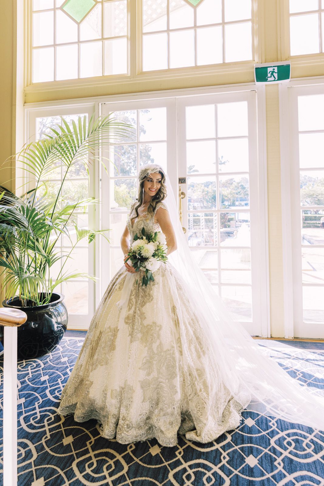 A bride poses in a flowing lace ball gown with cathedral train against large windows and tropical plants.
