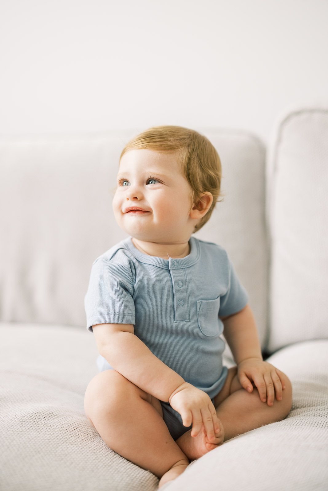 Baby in light blue onesie sitting on white couch looking up with a smile.