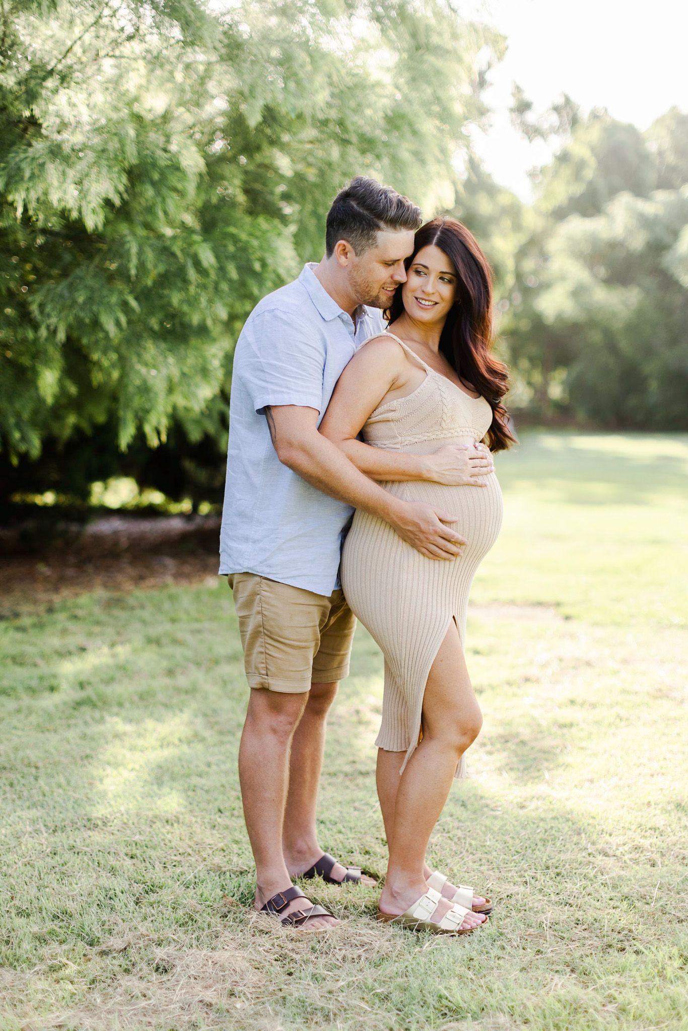 A couple embraces tenderly in a sun-drenched park setting during a maternity photo session.