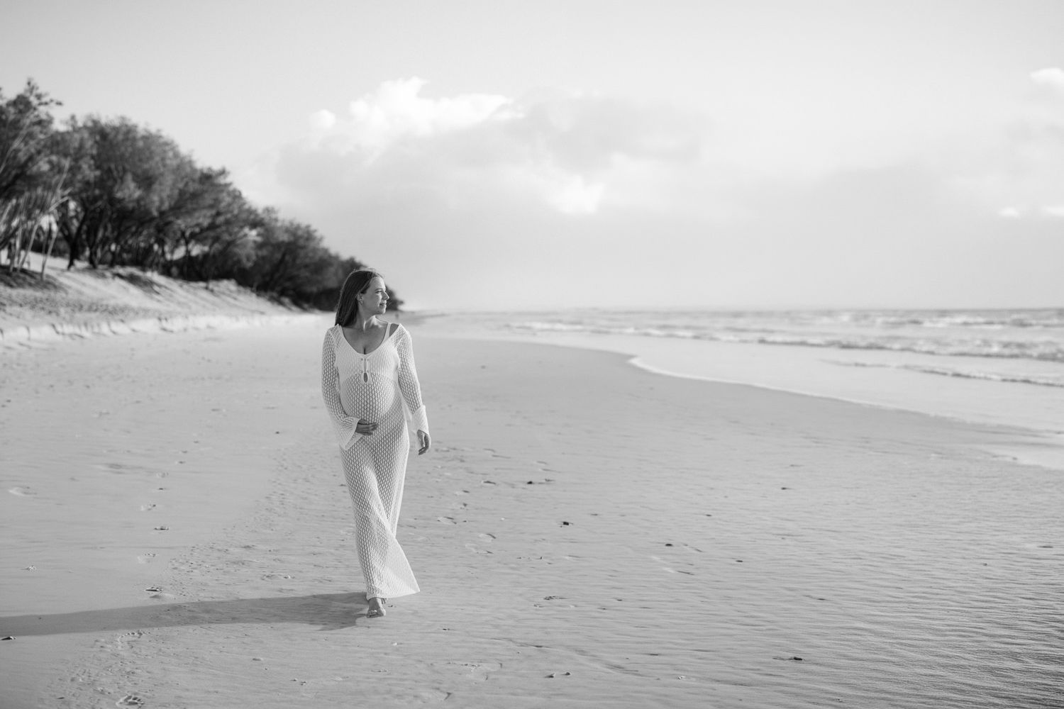 A woman in a white dress walks along a serene beach in this elegant black and white maternity photo.