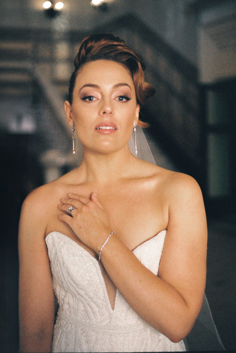 Elegant bride in strapless white gown with updo, diamond earrings, bracelet, and ring, posing gracefully indoors.