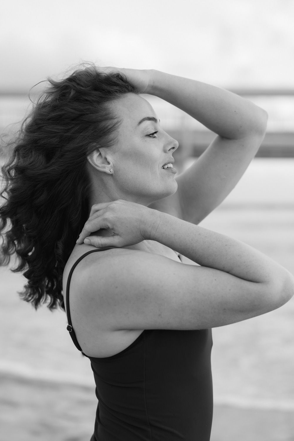 Black and white portrait of a woman with curly hair posing on a beach, hands raised gracefully.