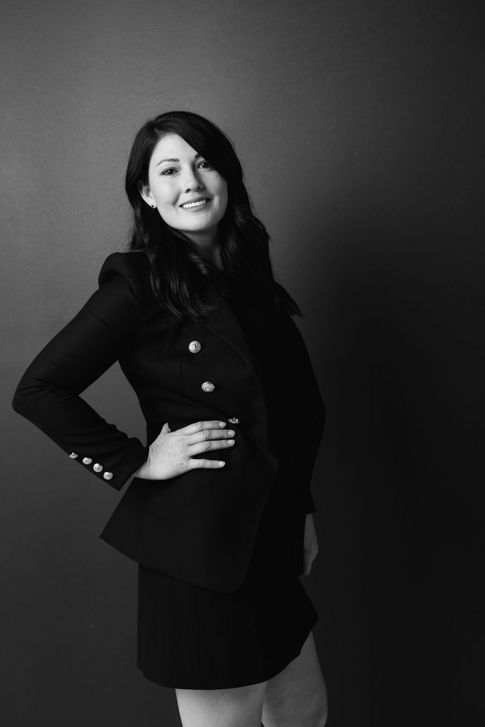 Black and white portrait of a woman in a tailored blazer with buttons posing against a dark background.