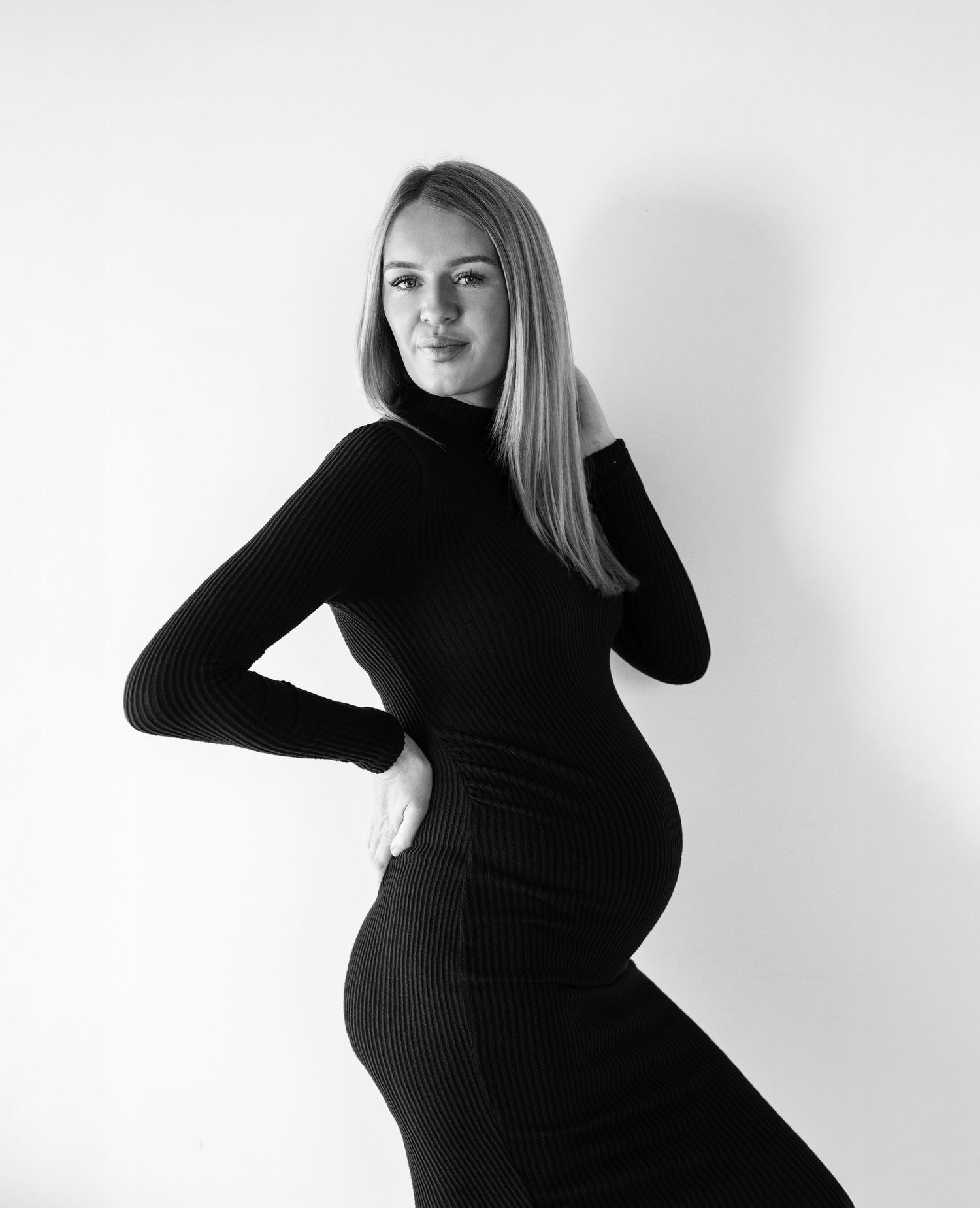 Side profile maternity portraits in black ribbed dress against white backdrop.