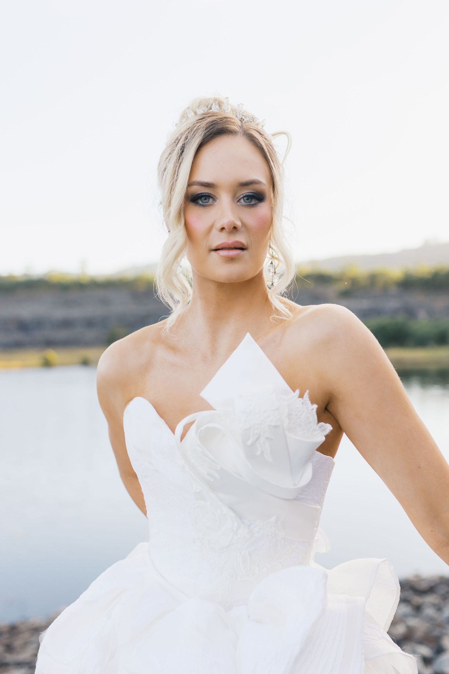 Bride in flowing white gown poses elegantly by waterfront during sunset photoshoot.