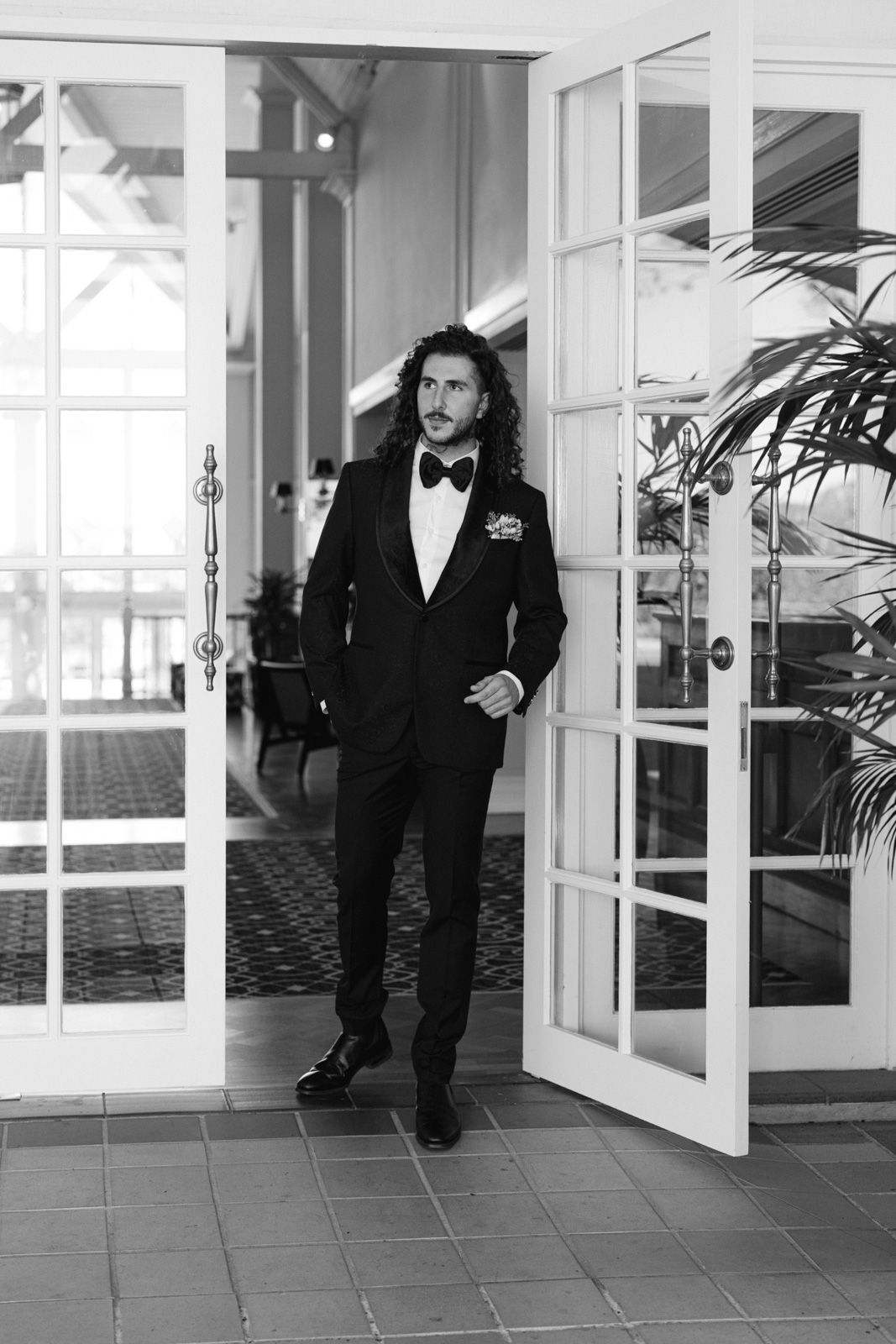 A person in a black tuxedo walks through an elegant hallway with white French doors and tiled floors.