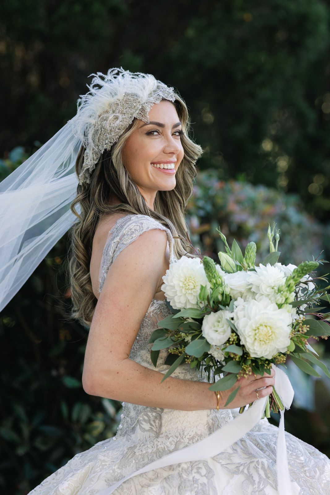 Beautiful bride in lace wedding dress and veil holds elegant white peony bouquet while smiling outdoors.