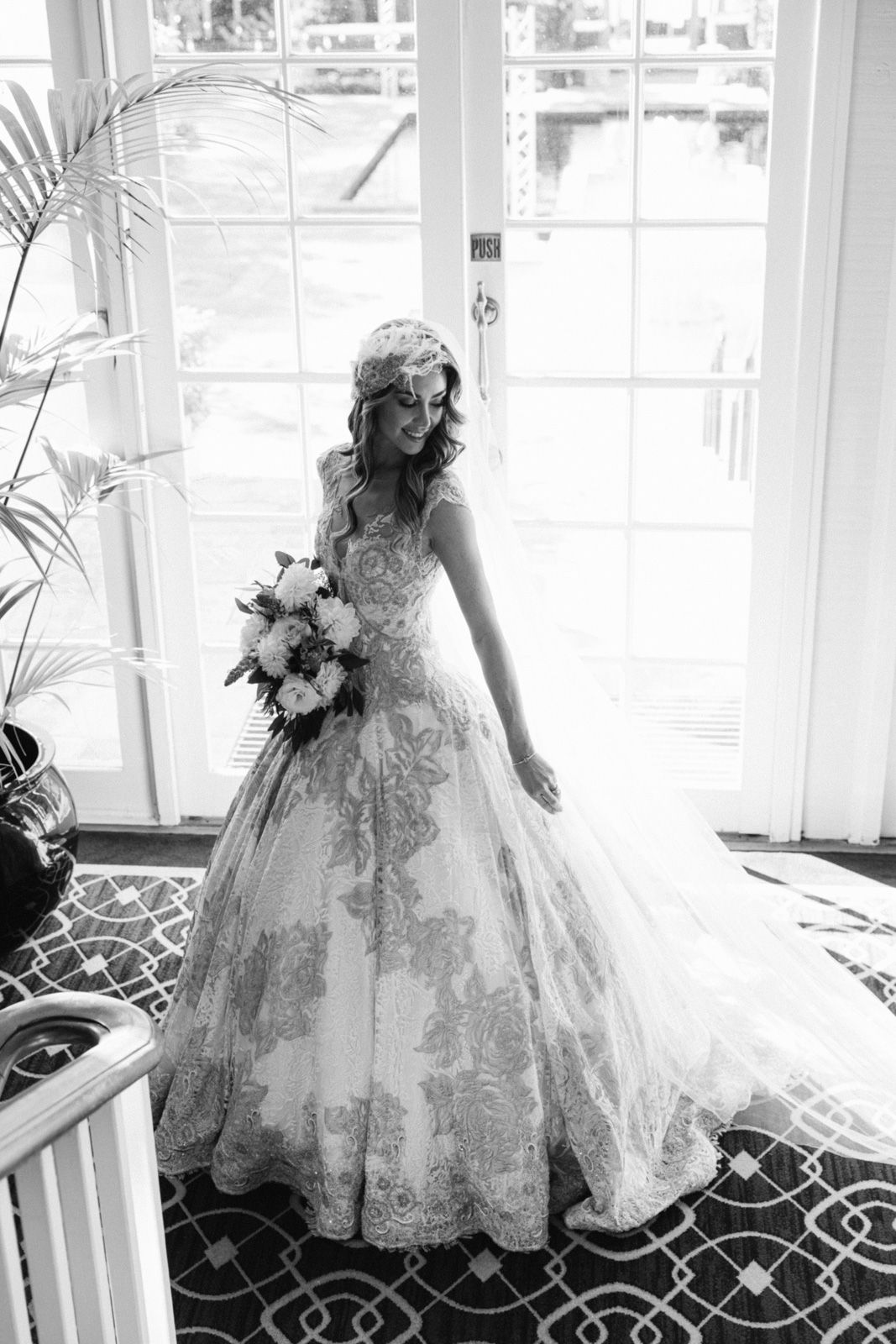 Black and white photo of a bride in an ornate lace wedding gown holding a bouquet while standing by French doors.