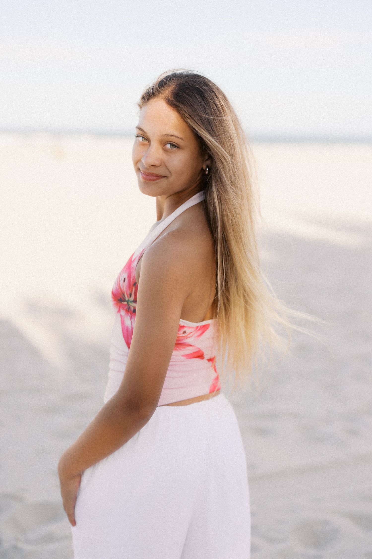 Young woman with long blonde hair wearing a floral crop top and white pants posing on a sandy beach.