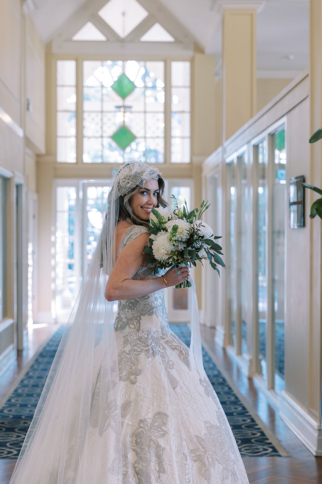 A bride in an elegant lace wedding gown holds a white and green bouquet in a bright hallway with decorative windows.