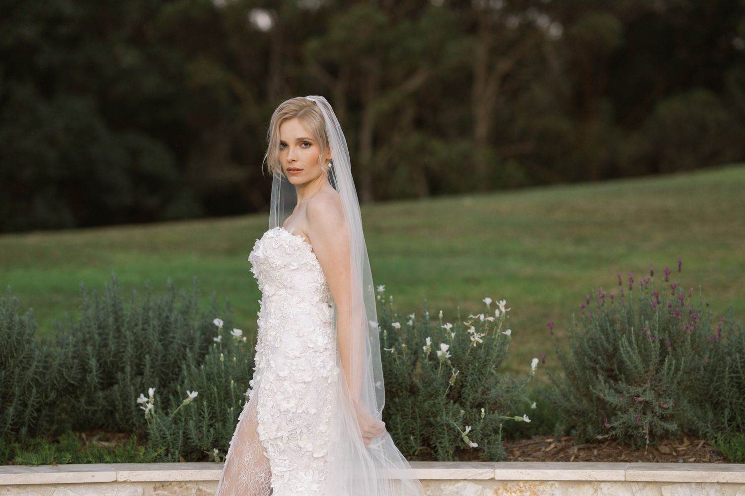 A bride in a white lace wedding dress poses in a garden setting with flowers and greenery in the background.
