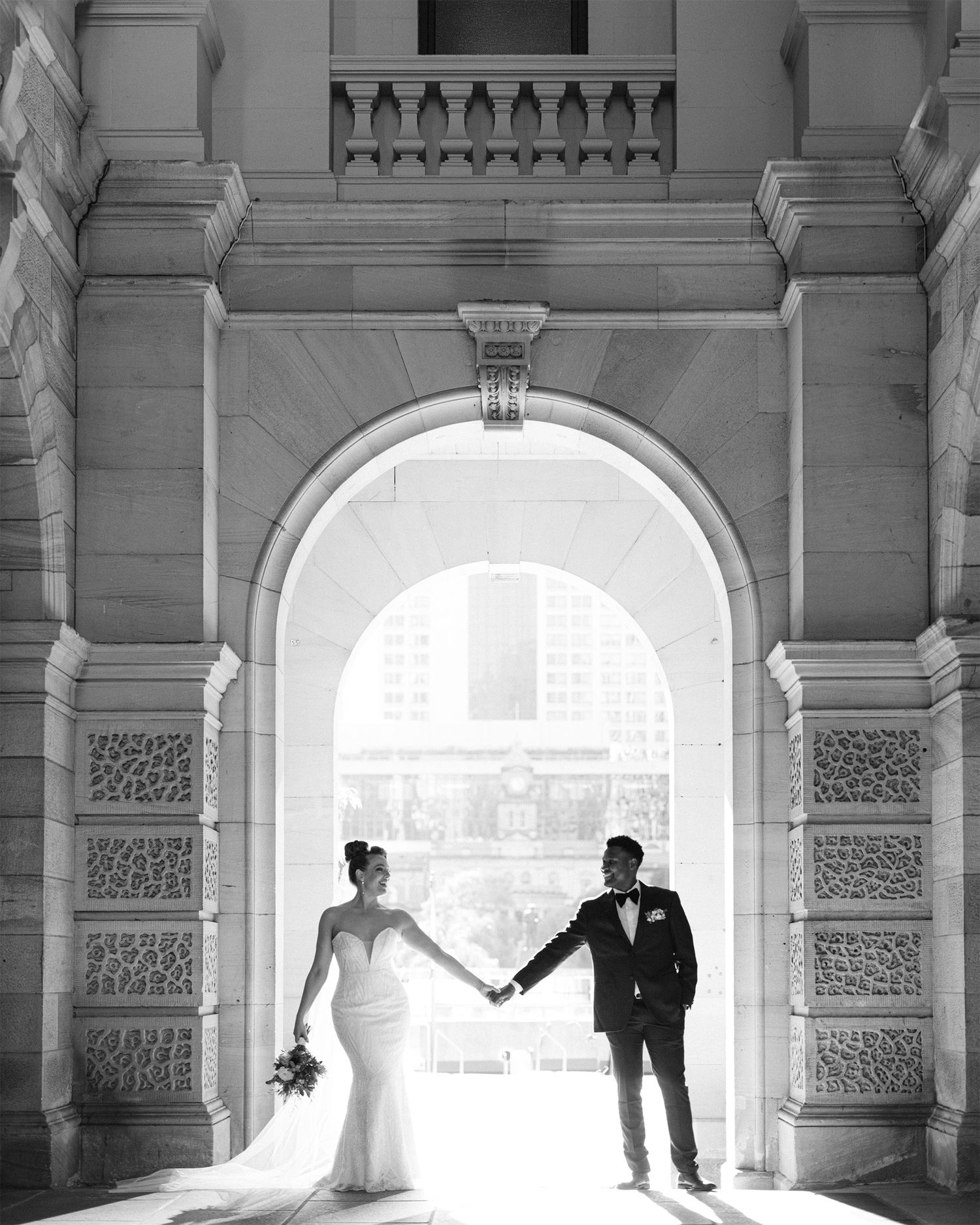 A dramatic black and white wedding photo of a couple silhouetted in a grand stone archway.