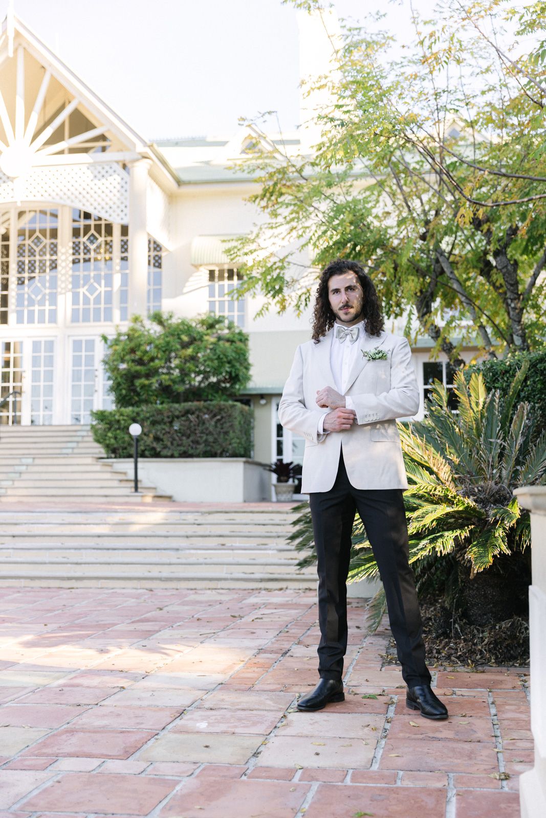 Elegant figure in white dinner jacket and black pants stands on brick patio outside grand building with ornate windows.