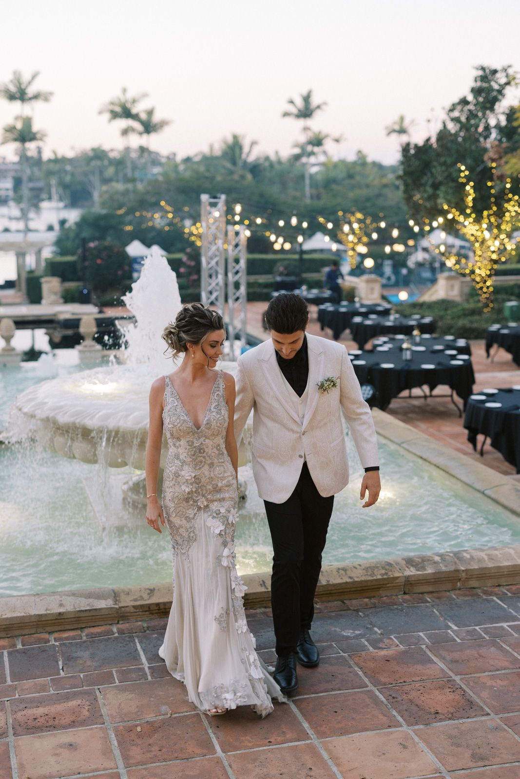 A couple in wedding attire walk together by an illuminated fountain at dusk with string lights and palm trees in background.