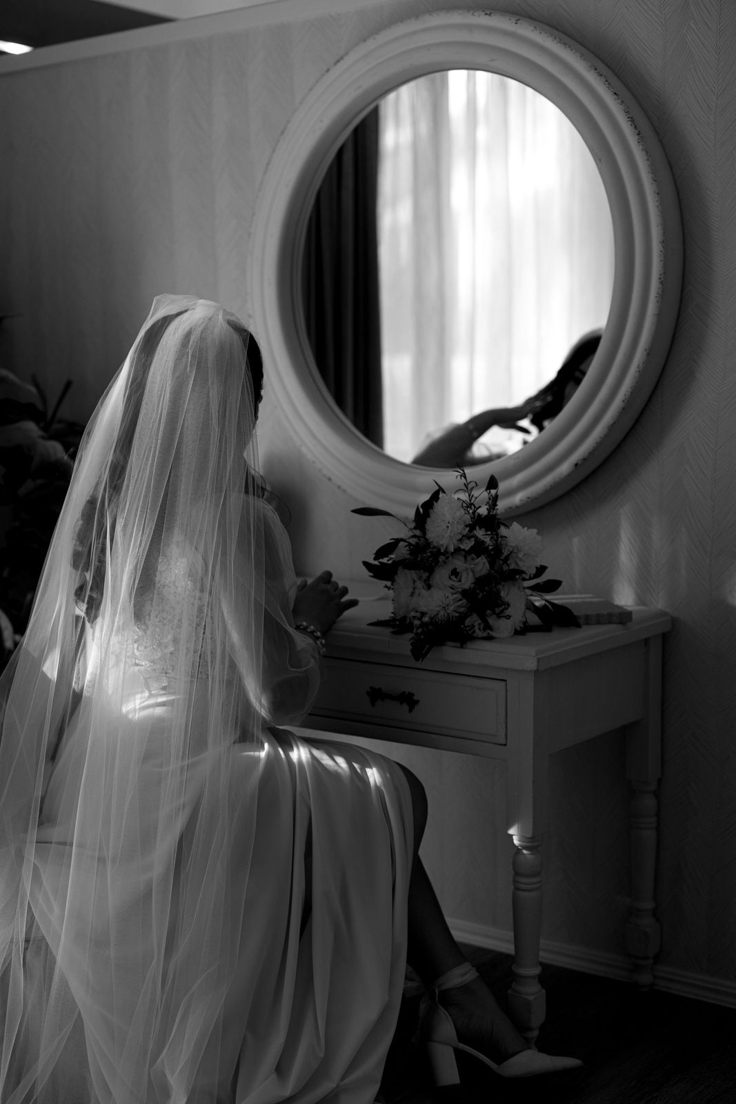A black and white photograph showing a bride in a flowing veil seated at a vanity with a round mirror and flowers.