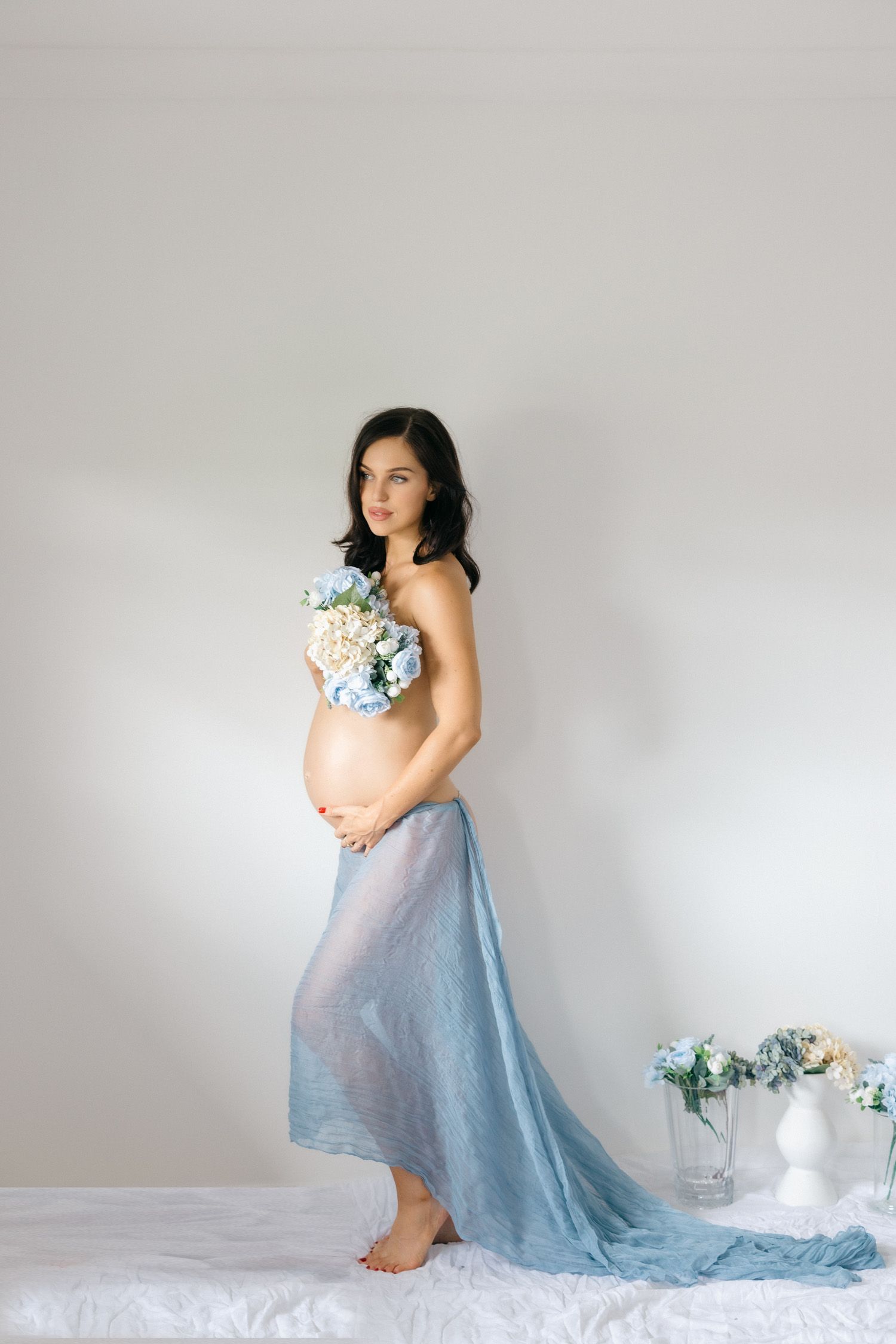 Pregnant woman in blue sheer skirt holding flowers, posing gracefully in a bright studio setting.