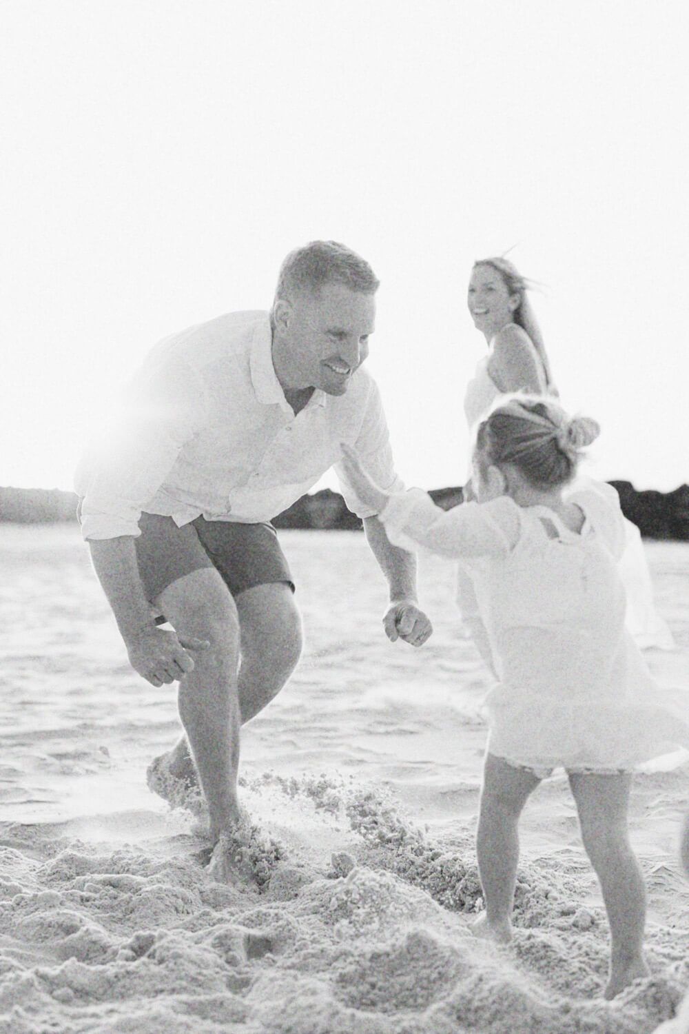 Black and white photo of a man playing with a young child on a sandy beach while a woman watches in the background.