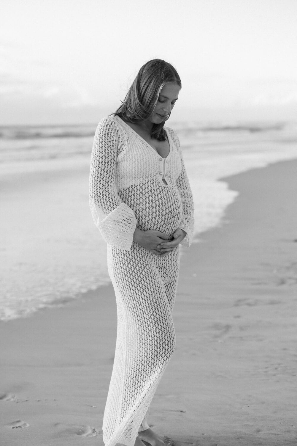 Pregnant woman in white crochet dress cradling her baby bump on a serene beach in black and white.