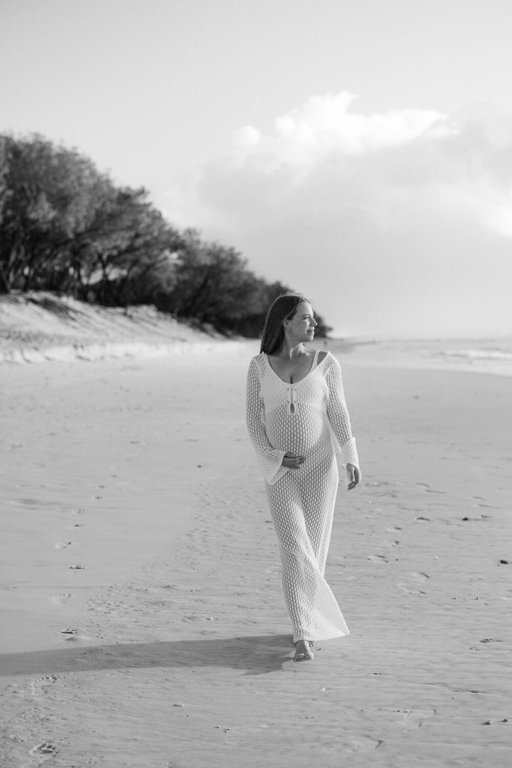 Pregnant woman in white mesh dress walking barefoot on sandy beach in black and white maternity photo.