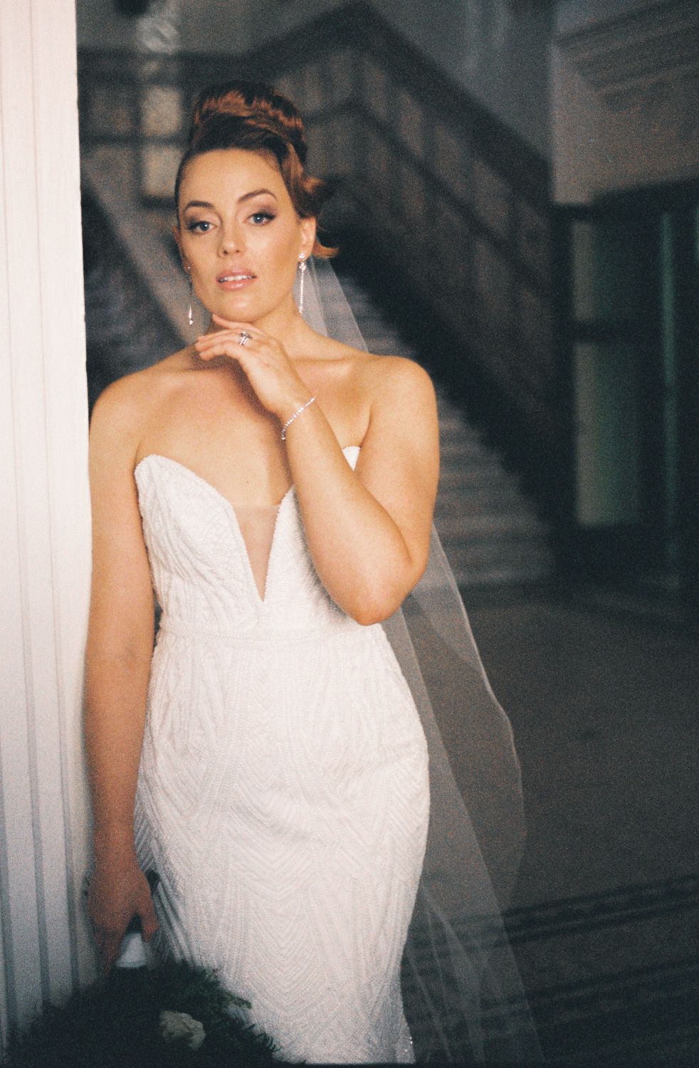 Bride in a strapless beaded white wedding gown with veil poses elegantly by a column in a grand venue.