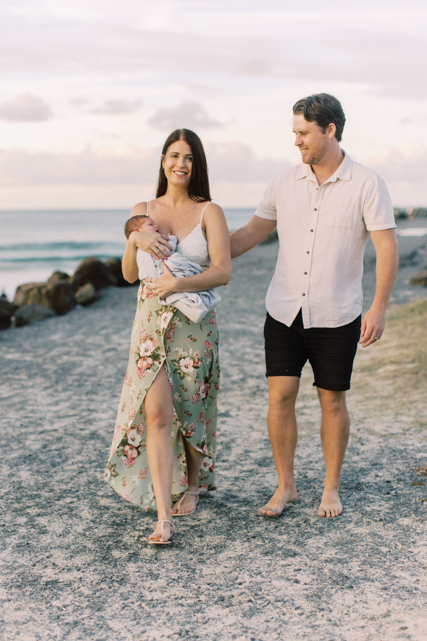 A family walks together along a rocky beach at sunset, wearing casual summer attire and enjoying the ocean view.