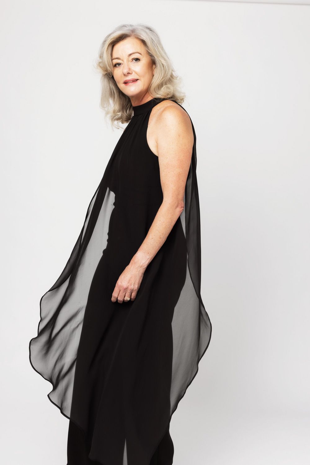 A model in an elegant black evening dress with sheer flowing panels poses against a white studio background.