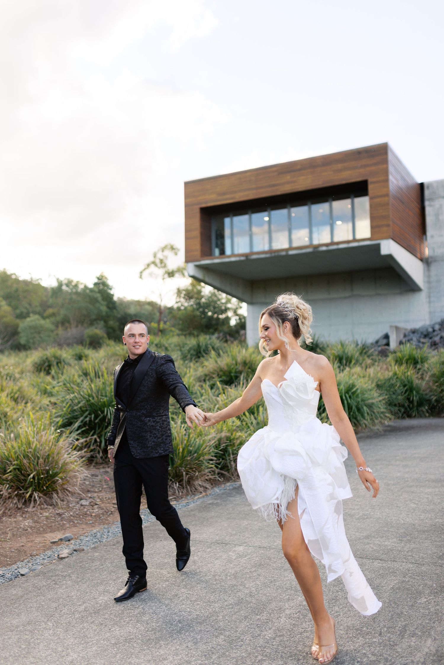 Wedding couple playfully running outside modern architectural building with mountain backdrop.