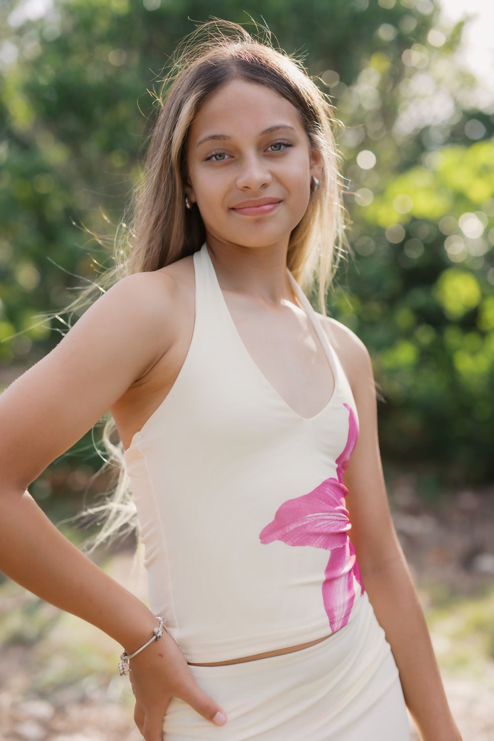 Young woman in cream halter top with pink floral print posing outdoors among green trees.