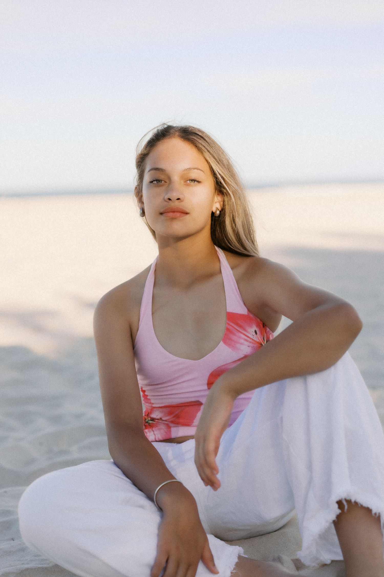 Young woman in pink floral halter top and white pants sitting on sandy beach, posing confidently in soft golden light.
