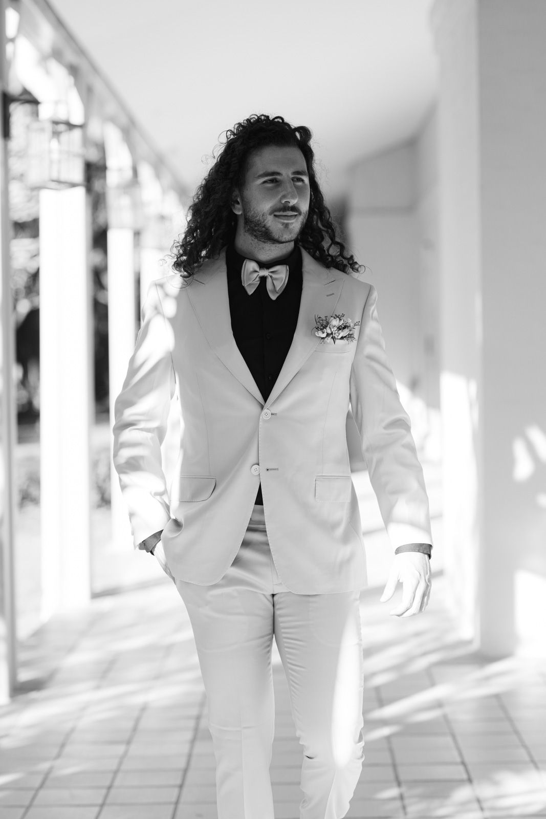 A black and white photo of a groom in a white tuxedo walking along a covered outdoor walkway.