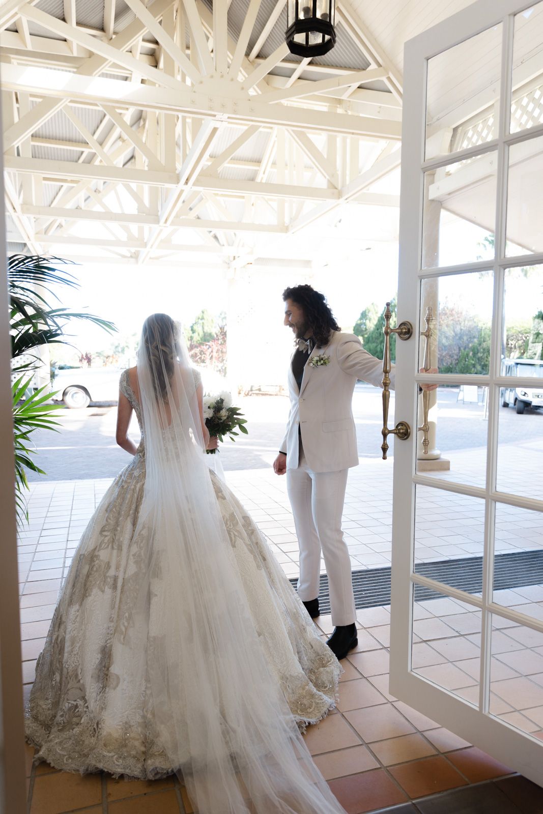 Multiple brides in white wedding gowns walk through a bright sunlit corridor with terracotta floors and glass walls.